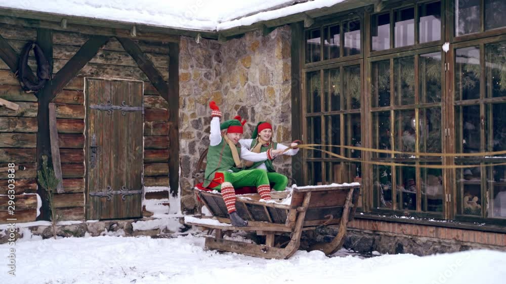 Christmas elves ride wooden sledges. New Year is coming. Christmas eve. Boy and girl wear christmas clothes. Having fun.
