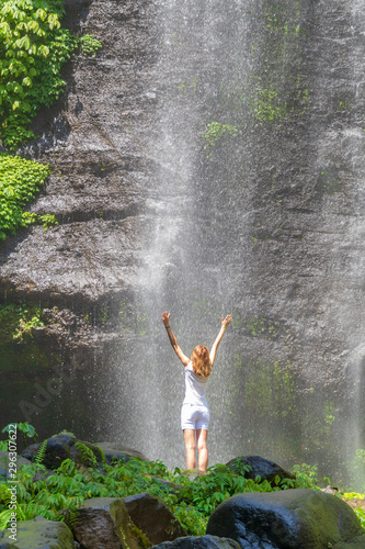 A young woman stands with her back in white clothes at the Sekumpul Great Waterfall in the deep rainforest of Bali Island, Indonesia.