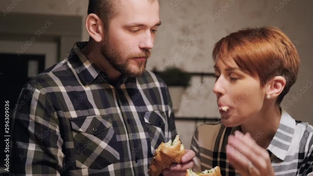 Young married couple in casual clothes, man and woman eating sandwiches at home in the kitchen. Evening light from the yellow lamps. Homeliness. Young married couple in casual clothes, man and woman eating sandwiches at home in the kitchen. Evening light from the yellow lamps. Homeliness.