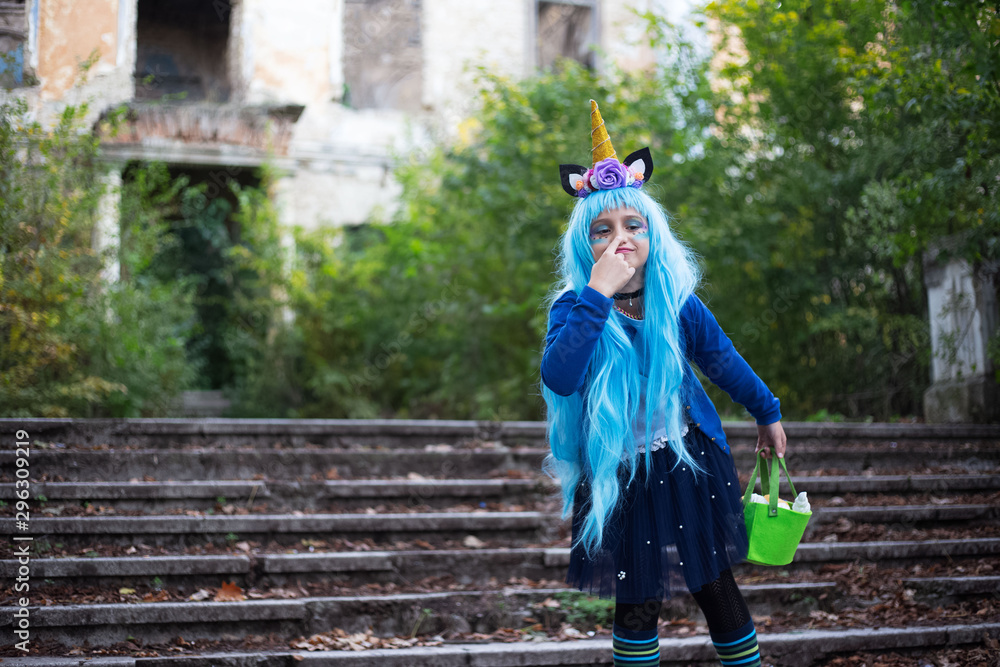Cheerful child girl with blue hair, touch nose with finger, wearing unicorn costume for halloween with orange horn. Holding green sweets bucket. Standing on stairs of scary abandoned house.