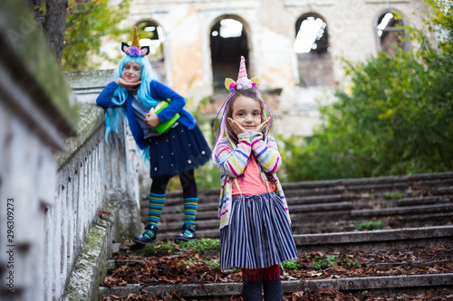 Two cheerful little girls  in colorful costume of unicorn  blue hair and green halloween bucket  standing on stairs of scary abandoned house.