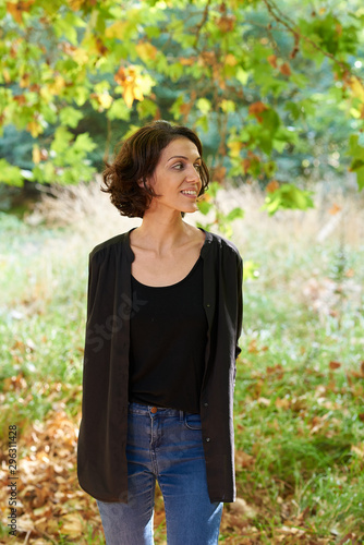 Latin brunette woman with a black t-shirt in a forest with dry leaves and autumnal colors, the sun enters between the branches of the trees.