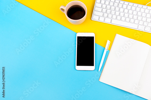 Flat lay design of office desk mock up with smartphone, labtop, notebook, pen and cup of coffee on two tone (yellow and blue) background with copy space. Top view. Work space and business concept.