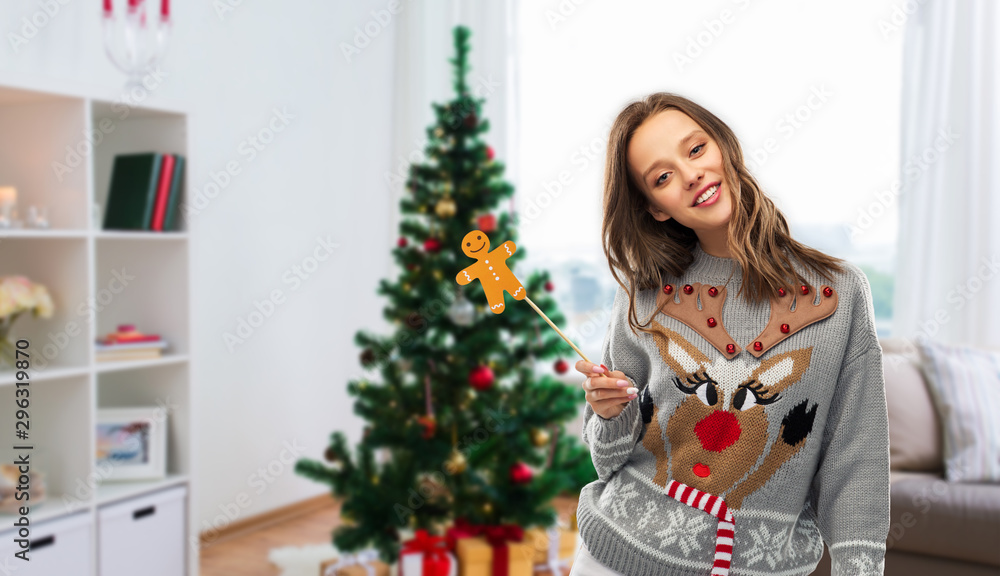 people and holidays concept - happy young woman in jumper with reindeer pattern holding gingerbread accessory at ugly sweater party over christmas tree on home background people and holidays concept - happy young woman in jumper with reindeer pattern holding gingerbread accessory at ugly sweater party over christmas tree on home background