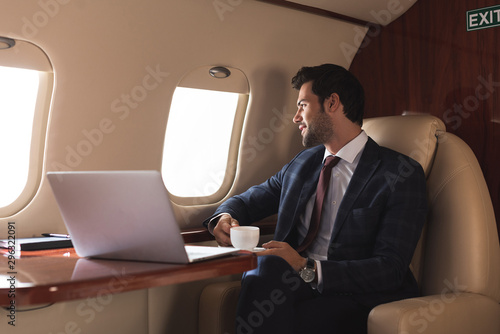 Obraz na plátne pensive businessman holding cup of coffee in plane with laptop during business t