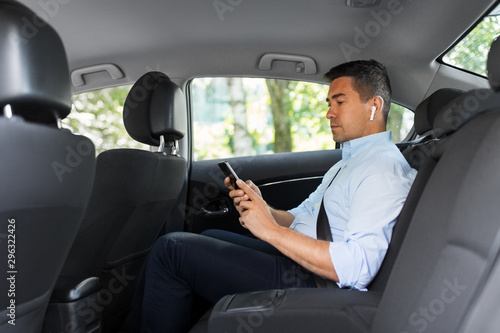 transport, business and technology concept - male passenger or businessman with wireless earphones using smartphone on back seat of taxi car