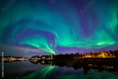Northern lights above lake. Green aurora on purple sky with stars and clouds. Trees  city light. Reflections in water. Prestvannet  Tromso  Norway.