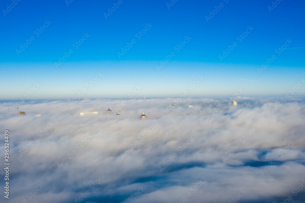 Quadrocopter flying over the fog at high altitude over the city. The upper floors of high-rise buildings among huge clubs of white fog with a horizon line on a crystal clear sky. Quadrocopter flying over the fog at high altitude over the city. The upper floors of high-rise buildings among huge clubs of white fog with a horizon line on a crystal clear sky.
