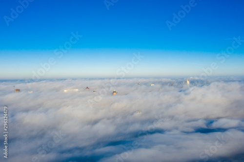 Quadrocopter flying over the fog at high altitude over the city. The upper floors of high-rise buildings among huge clubs of white fog with a horizon line on a crystal clear sky.