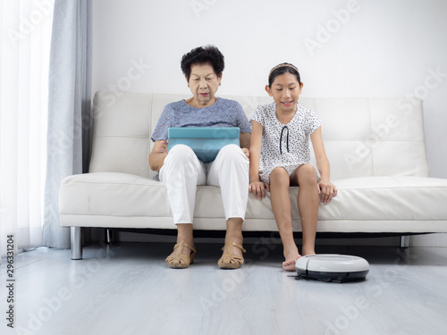 Asian senior woman and girl unsing tablet while robot vacuum cleaning floor at home. Modern lifestyle concept.
