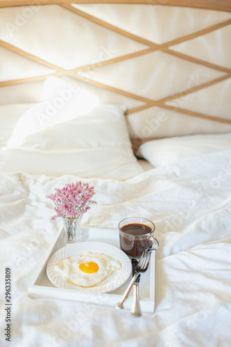 White tray with breakfast on a bed in a hotel room. Fried egg, cup of coffee and flowers in white sheets in light bedroom.