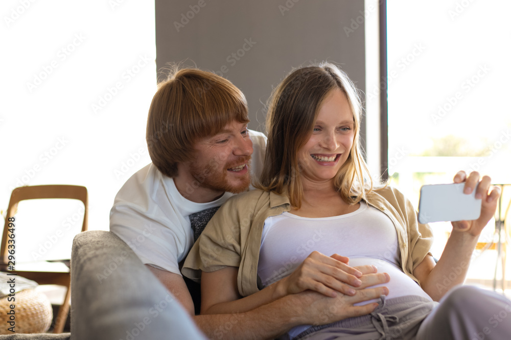 Joyful excited expectant parents using smart phone for selfie or video  call. Man embracing pregnant wife lying on couch, holding cellphone,  smiling, laughing. Funny video talk concept Stock Photo | Adobe Stock