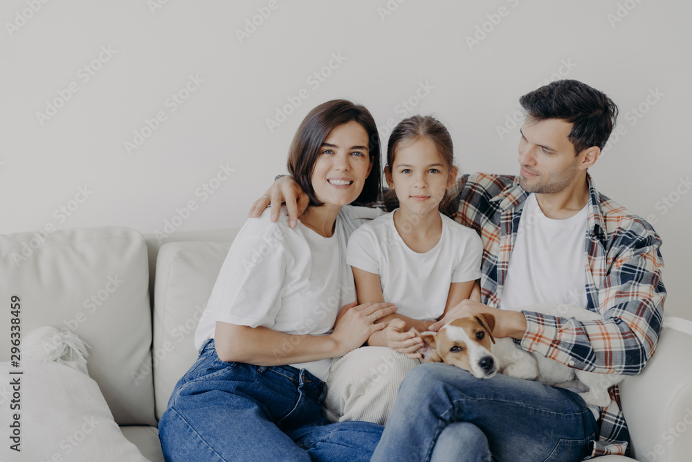 Family time concept. Cheerful mum, dad and small girl embrace all together, spend spare time in living room of their new home, sit on sofa, play with dog, have perfect relationships, enjoy comfort Family time concept. Cheerful mum, dad and small girl embrace all together, spend spare time in living room of their new home, sit on sofa, play with dog, have perfect relationships, enjoy comfort