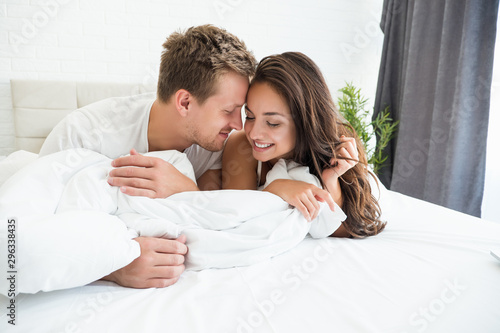 young couple handsome man and beautiful woman lying in bed in bright bedroom feeling love