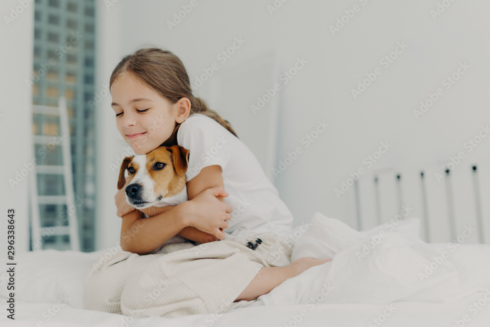 Pleased little girl plays with pet, embraces dog and keeps eyes closed from pleasure, dressed in nightclothes, poses on bed after waking up, expresses love to pedigree animal. Bedding concept Pleased little girl plays with pet, embraces dog and keeps eyes closed from pleasure, dressed in nightclothes, poses on bed after waking up, expresses love to pedigree animal. Bedding concept