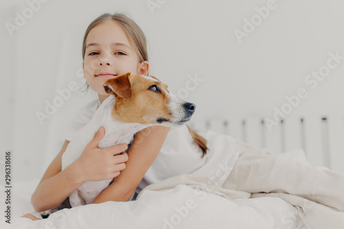 Little girl embraces small pedigree dog, stay in bed, plays with favourite pet before sleep, has appealing look, gazes camly into camera. Childhood and bed time concept. Kid hugs domestic animal photo