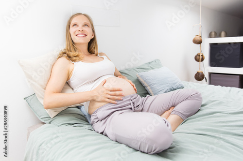 Cheerful pregnant woman relaxing at home. Happy beautiful young woman sitting on bed, touching belly, looking up, smiling. Expecting child concept