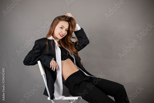 Studio portrait of a beautiful girl in a strict business suit, sitting on a chair on a gray background. Business woman. Sexy girl with red hair