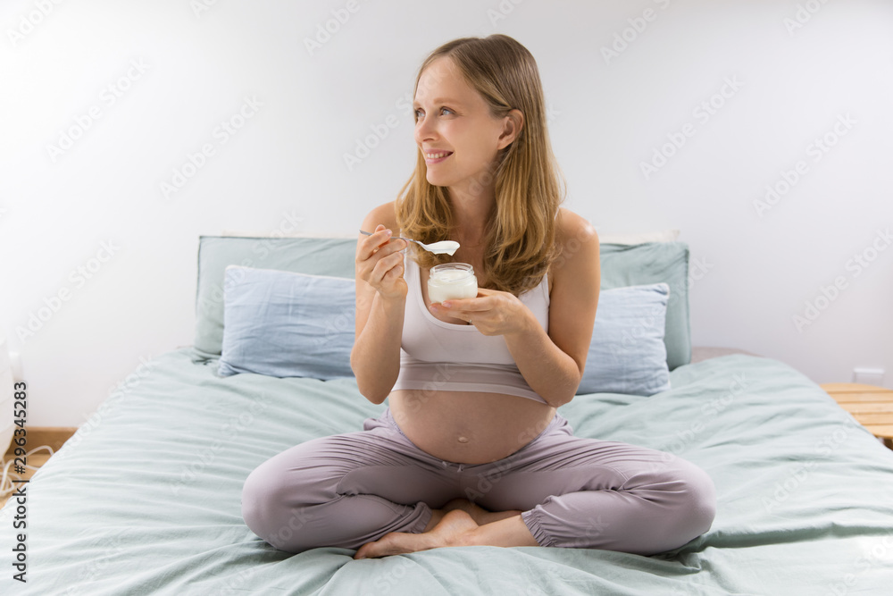 Joyful expectant mother enjoying healthy food. Pregnant woman sitting on bed, eating yogurt, looking away, smiling. Pregnancy and healthy meal concept Joyful expectant mother enjoying healthy food. Pregnant woman sitting on bed, eating yogurt, looking away, smiling. Pregnancy and healthy meal concept