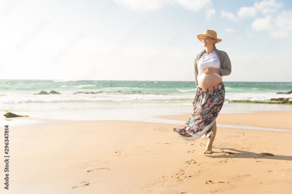 Relaxed pregnant woman holding hand on belly. Cheerful future mother walking on beach during vacation. Pregnancy concept Relaxed pregnant woman holding hand on belly. Cheerful future mother walking on beach during vacation. Pregnancy concept