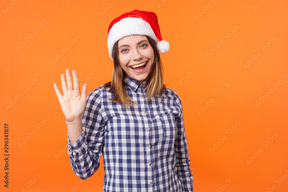 Hello! Portrait of friendly kind brunette woman in santa hat and checkered shirt waving raised hand and saying hi to camera, enjoying christmas time. indoor studio shot isolated on orange background Hello! Portrait of friendly kind brunette woman in santa hat and checkered shirt waving raised hand and saying hi to camera, enjoying christmas time. indoor studio shot isolated on orange background
