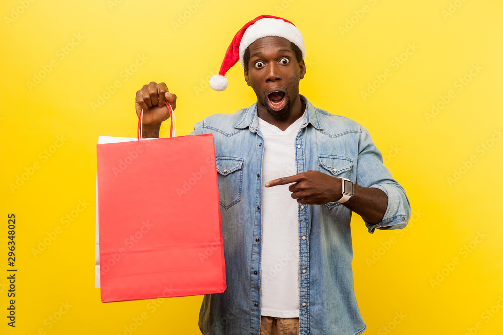 Portrait of surprised handsome young man in santa hat and casual denim shirt standing, pointing at shopping bag, looking shocked about xmas discounts. indoor studio shot isolated on yellow background