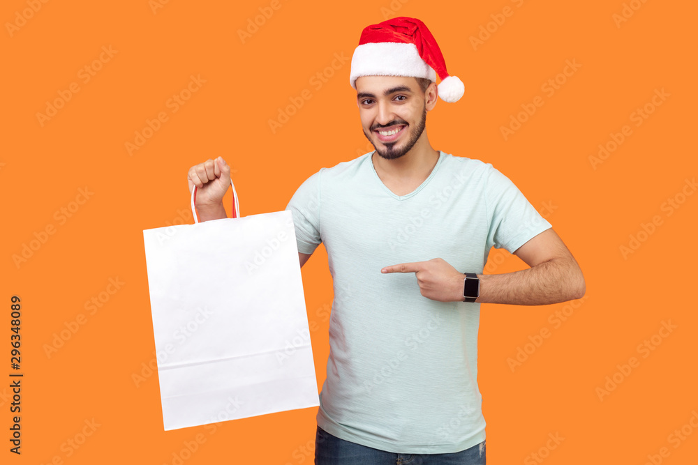 Portrait of cheerful bearded young man in santa hat and casual white t-shirt standing pointing at shopping bag and looking at camera with toothy smile. indoor studio shot isolated on orange background