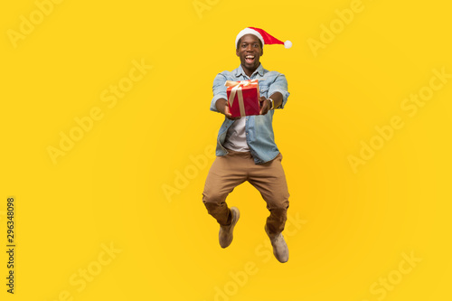 Full length portrait of extremely happy young man in santa hat and casual denim shirt jumping or flying and giving christmas gift box to camera. indoor studio shot isolated on yellow background
