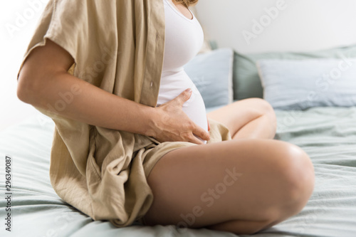 Expectant mother sitting on bed and holding tummy. Pregnant woman relaxing at home and touching belly. Expecting baby concept