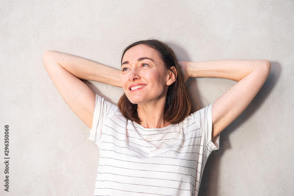 Close up older woman smiling with hands behind head against white wall Close up older woman smiling with hands behind head against white wall