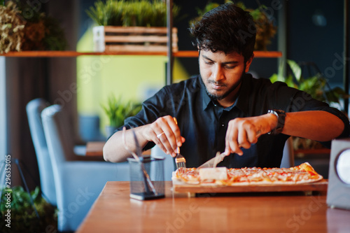 Confident young indian man in black shirt sitting at pizzeria with pizza.