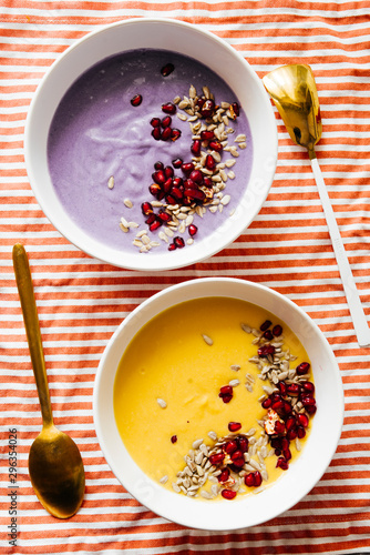 Pumpkin and coconut soup with pumpkin seeds and pomegranate and the other violet potatoes soup, on a colored striped towel,flat lay composition, healthy warm food for autumnal and winter season.