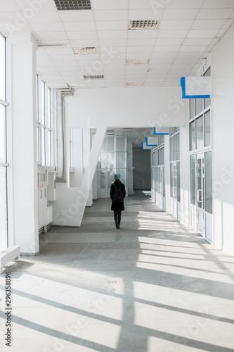 Back view of young pretty woman with short blue hair in black coat walking through the long white corridor of the building with many windows and doors. © Artem