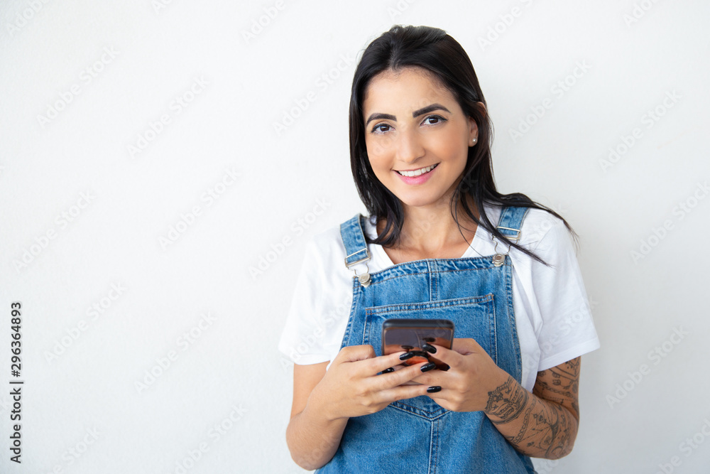 Front view of smiling young woman texting on smartphone. Cheerful brunette holding smartphone and looking at camera. Technology concept Front view of smiling young woman texting on smartphone. Cheerful brunette holding smartphone and looking at camera. Technology concept