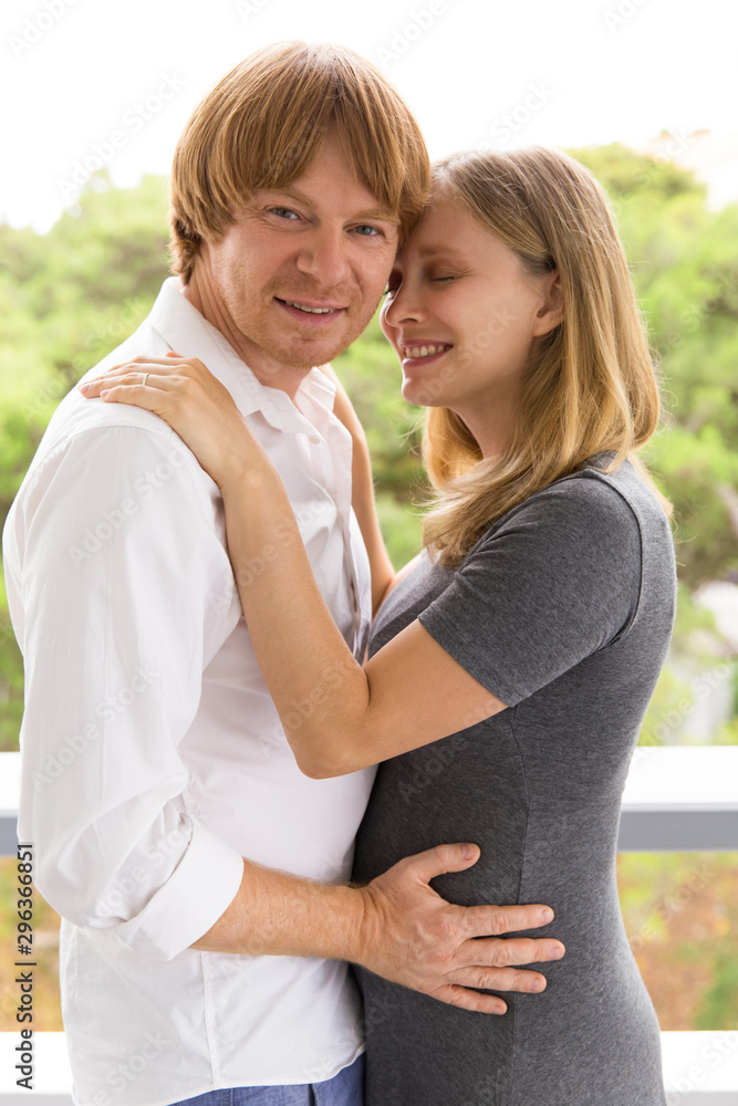 Fototapeta premium Happy expectant parents young posing on terrace. Man and pregnant woman standing outside, hugging each other, talking, looking at camera. Happy family couple concept