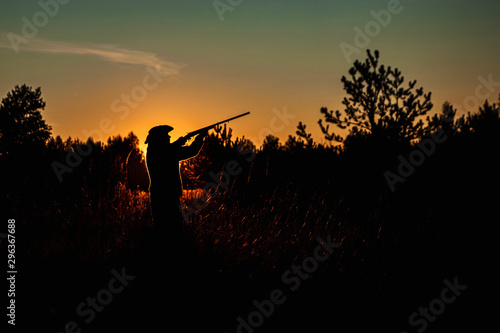 Silhouette of a hunter in a cowboy hat with a gun in his hands on a background of a beautiful sunset. The hunting period, the fall season is open, the search for prey. © Aliaksandr Marko