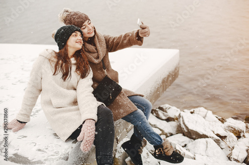 two bright and merry girls sitting in the frozen snowy park neaw water and use the phone photo