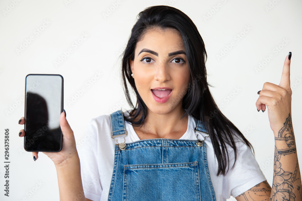 Young woman with open mouth holding smartphone. Front view of cheerful brunette showing smartphone and pointing up with finger. Technology concept Young woman with open mouth holding smartphone. Front view of cheerful brunette showing smartphone and pointing up with finger. Technology concept