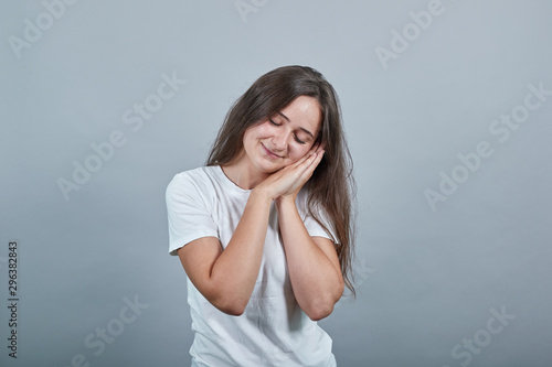 Young caucasian girl with white t-shirt making sleep gesture in dorable expression. On a grey background in the Studio she put her two hands together and rested her head on them