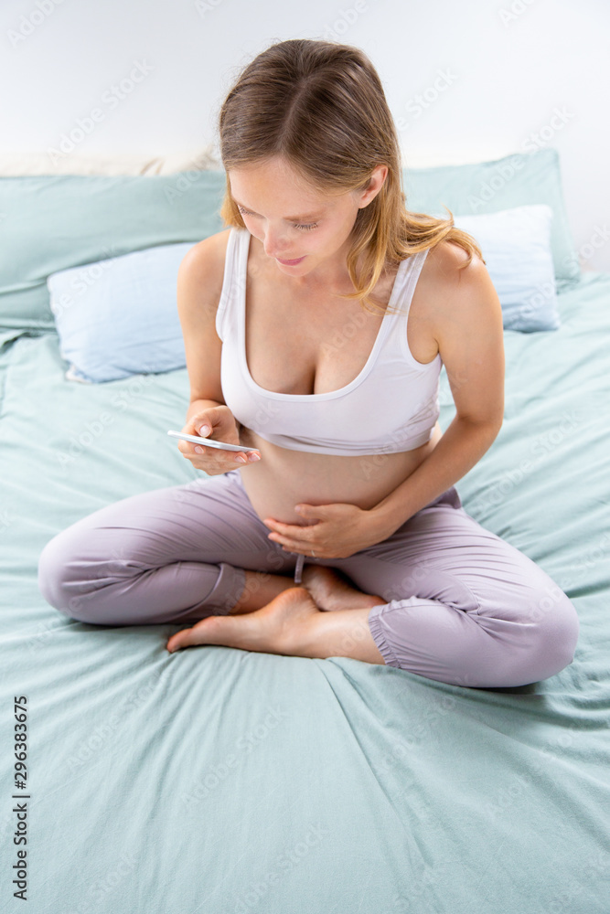 Focused expectant mom using online mobile app on smart phone. Pregnant woman sitting on bed, using cellphone, typing, looking at screen. Wi-Fi concept Focused expectant mom using online mobile app on smart phone. Pregnant woman sitting on bed, using cellphone, typing, looking at screen. Wi-Fi concept