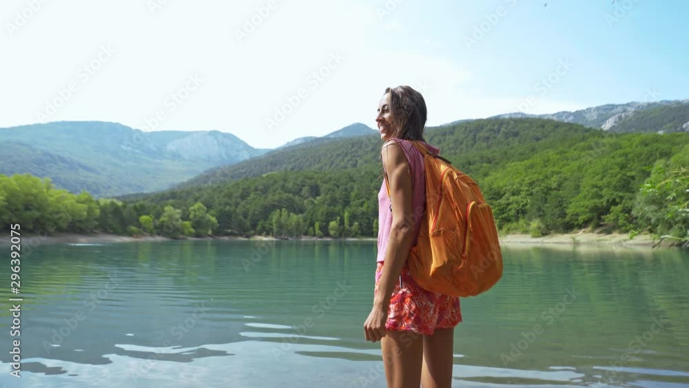 Woman hiker enjoying a landscape with mountain lake. back view of smiling young slender woman with small orange backpack standing on mountain lake background with rising hands Woman hiker enjoying a landscape with mountain lake. back view of smiling young slender woman with small orange backpack standing on mountain lake background with rising hands