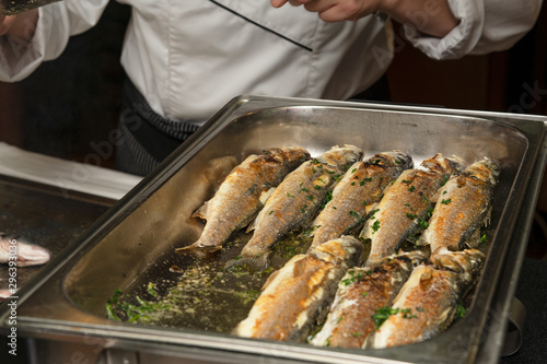 The restaurant in the hotel. Buffet table. The cook roasts and spreads fried fish on a baking sheet. The cook is shown in fragments. Selective focus.