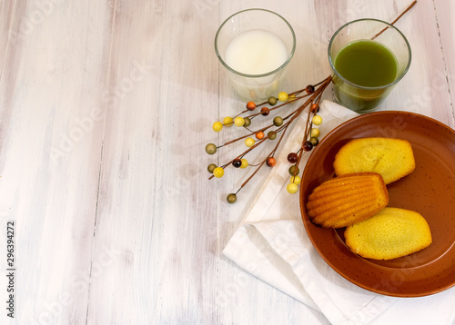 Top view of madeleines, matcha tea and milk over orange plate in wooden rustic background with copy space 