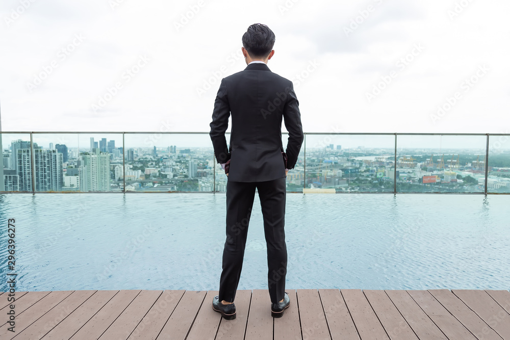 Asian businessman in a black suit is standing on a wooden floor by the pool on the roof with blurred of the building background.
