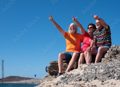 Senior caucasian people sitting on the cliffs smiling with happiness. Three friends enjoying free time and retirement. Blue sky, wind and kitesurf. Arid mountain