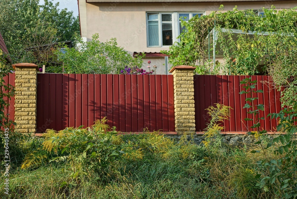 fence of red metal and brown bricks overgrown with green vegetation and grass fence of red metal and brown bricks overgrown with green vegetation and grass