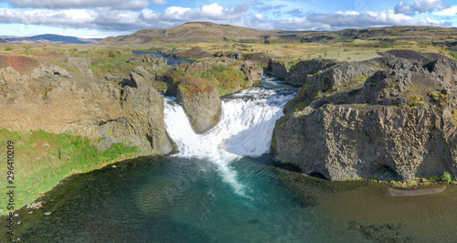 Epic aerial drone view flying over landscape of Hjalparfoss waterfall and lagoon on a sunny day.