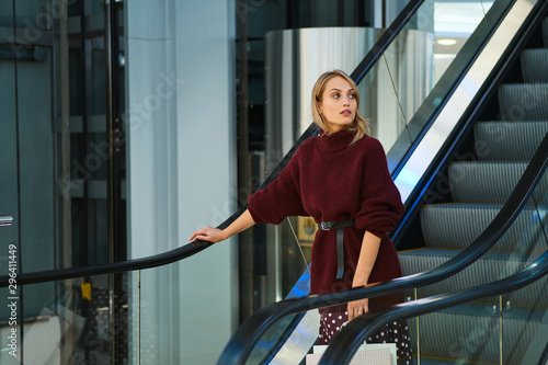 Beautiful blond girl with shopping bags on escalator dreamily looking away in shopping mall