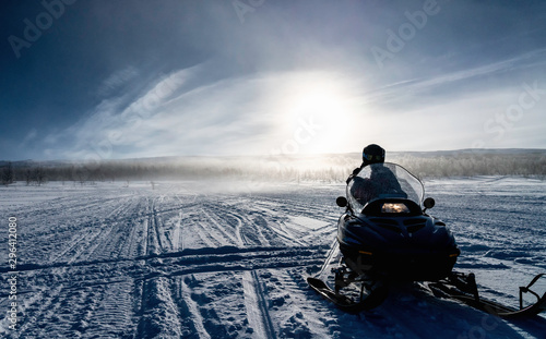 One person ready to drive snowmobile in very cold mountains in Sweden, frosty fog around bright sun creates halo effect, birches forest and mountains behind driver. Hemavan -Tarnaby area, Lappland