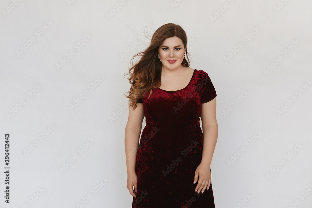 Beautiful plus size model girl in modish red velvet dress isolated at white background. Young fat woman with bright makeup and with stylish hairstyle posing in studio. Concept of XXXL fashion